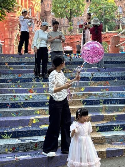 A group of people are positioned on a colorful mosaic staircase in the city of Baqiao, China. The staircase is decorated with tile mosaics of flora and fauna. A girl in a white dress and a hair clip looks at a person in a striped shirt holding a pink glitter balloon on a stick. Several other people are on the staircase, including a woman with a sunhat, and two men. In the background, an ornate red brick building is visible.