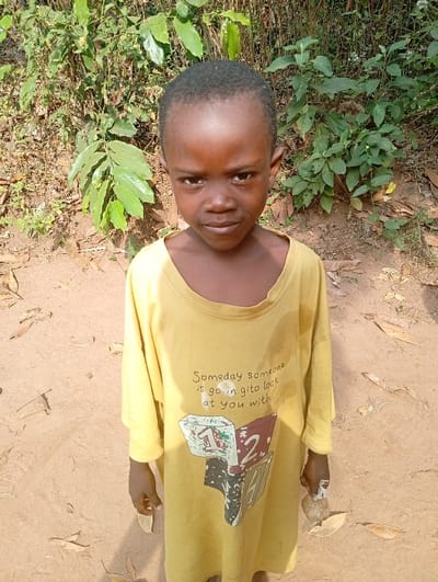 A young child stands facing the camera in an unpaved outdoor area of Eziudo, Nigeria. The child has dark skin and short, dark hair, looking directly forward with a neutral expression. They wear an oversized, long-sleeved, yellow t-shirt with text printed on the front that reads, "Someday someone is go in gito lock at you with," above graphics depicting stacked, geometric blocks with numbers "1" and "2." The t-shirt appears stained around the collar and chest. In the child's right hand, a small, irregularly shaped light-colored object, possibly wrapped in clear plastic, is held. The ground beneath the child is light brown, sandy earth, sparsely covered with scattered dry leaves. The background consists of dense green foliage and various bushes, indicative of natural vegetation.