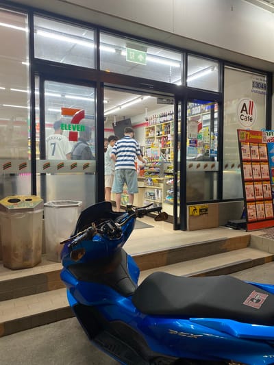 This media file captures an outdoor scene at a 7-Eleven convenience store in Baan Bo Phut, Thailand, likely at night given the artificial lighting. A bright blue scooter is parked in the foreground on the right, its seat prominently displaying a logo that reads "NA THONG."
The storefront is visible, with large glass windows revealing the interior. Inside, shelves stocked with various products are illuminated. Several people are visible through the glass. One man, wearing a striped t-shirt and light-colored shorts, stands near the entrance. Another individual, wearing a white shirt with the number "7" and "BECKHAM" printed on it, is also seen inside. A "7-ELEVEN" logo is clearly visible on the store's exterior.
Text elements within the image include the "7-ELEVEN" branding, "All Café" signage, and Thai text which includes "CPALL บมจ. ซีพี ออลล์" and "13048 บ้านดอนสมุย" (likely identifying the company and the specific store location). There are also yellow warning signs indicating a change in floor level ("พื้นต่างระดับ" and "YOUR DEP"). The overall atmosphere appears to be that of a typical convenience store setting, with the presence of the scooter suggesting it's a common mode of transportation in the area.
