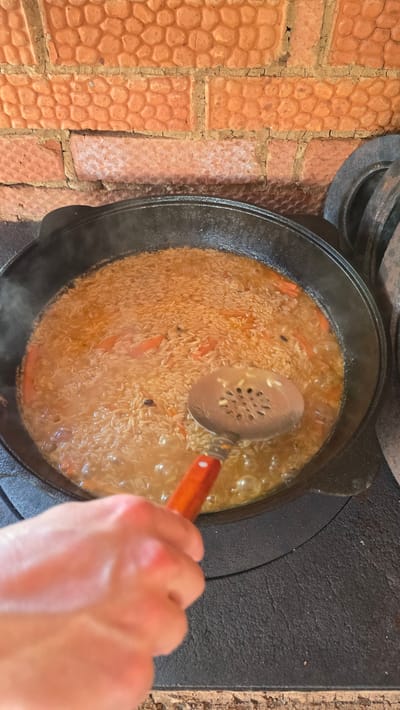 In the city of Забегалово, Russia, a close-up, elevated perspective shows a large, dark cast-iron kazan pot centrally positioned over a stove. The pot contains a light brown rice dish, intermingled with distinct orange carrot pieces and dark spices, simmering in a reddish-orange liquid, actively bubbling with visible steam rising. A fair-skinned human hand is visible in the lower-left, holding a metallic slotted spoon with a red-orange wooden handle, the spoon partially submerged in the contents of the pot. The kazan rests on a dark, textured, circular cast-iron stove surface; additional circular cast-iron rings are stacked on the stove to the right. The background features a rustic wall composed of irregularly shaped, reddish-orange bricks with visible, uneven mortar lines.