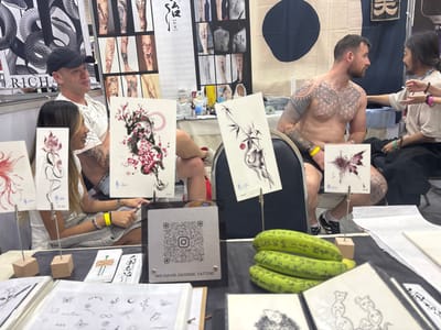 This image depicts an indoor scene at a tattoo convention or studio booth in Ban Rong Ruea Kham, Thailand. In the foreground, a display table showcases various tattoo designs. Prominently displayed are four upright samples: a red floral design, a black dragon amidst pink cherry blossoms, a bamboo motif with a red sun, and a purple butterfly. Below these, several flat sheets present smaller, intricate designs, including butterflies, flowers, and celestial symbols. A stand featuring a QR code linked to "INS:DAVID.GEORGE.TATTOO" is positioned centrally on the table. To the right, three green, elongated objects resembling gourds or cucumbers, inscribed with text, are stacked.
On the left side of the frame, a male wearing a black baseball cap and a white t-shirt is seated next to a female with long hair, dressed in a light-colored top. Both are observing the activity to their right. On the right side, a shirtless male with extensive tattoos covering his chest, arms, and legs is seated in a chair, looking towards another individual positioned further to the right. This individual, with long hair, wearing a light-colored top and dark pants, is facing the tattooed male, appearing to be engaged in a discussion or preparation for a tattoo session. The background features multiple banners, including a black and white snake design on the left and a cream-colored fabric banner with a dark circular design and Japanese characters on the right. Tattoo equipment and supplies are visible on a table behind the shirtless male.