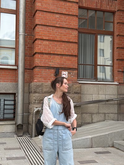 A young woman stands on an outdoor staircase, looking up and to the left. She has long, dark hair that falls over her shoulders and is wearing light blue denim overalls over a white long-sleeved shirt. A black bag with the word "MORGAN" in gold lettering is slung across her body.
The setting appears to be an urban environment, likely Ufa, Russia, given the context. The woman is positioned on a wide staircase made of grey stone tiles. To her left is a building with a facade of red brick, featuring a large window with dark wooden frames. Above the window, the brickwork has an interesting stepped design. A metal pipe runs horizontally across the brick wall, below the window.
The time of day is difficult to determine precisely, but the lighting suggests it could be daytime. There are no strong indications of the weather, though the absence of visible precipitation or extreme lighting conditions implies it is likely fair.
The woman's expression is one of looking upwards, perhaps with curiosity or wonder. Her posture is relaxed, with one hand resting on her hip. The overall mood is casual and everyday.
Visible text: "MORGAN" on the woman's bag. A "no smoking" sign is also visible on the brick wall to the left of the woman.