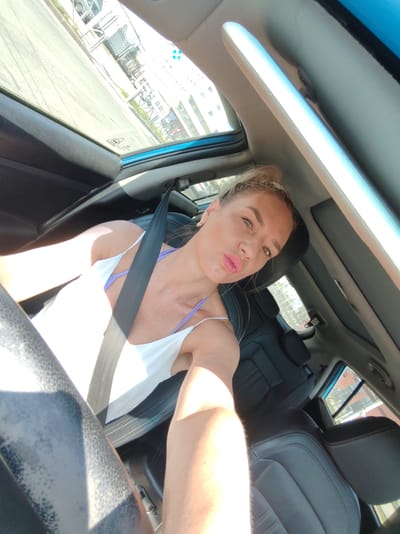 A woman is taking a selfie inside a car. She is wearing a white camisole with purple straps and a seatbelt is visible across her chest. Her hair is pulled up in a bun, and she is making a duck face. The interior of the car is visible, including black leather seats and a sunroof. Outside the car, a street and buildings are visible. The image appears to have been taken in Petropavlovsk-Kamchatsky, Russia.