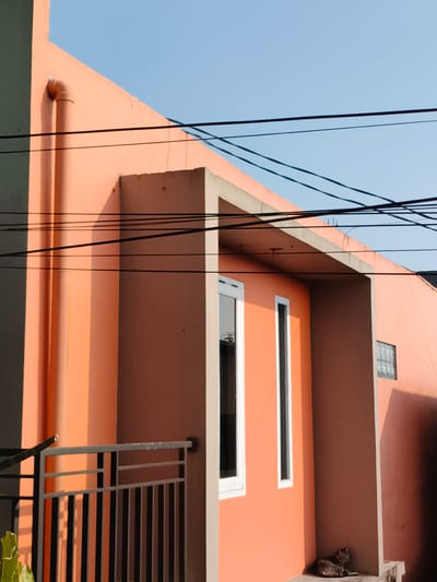 A salmon-colored building stands under a clear blue sky in Pacet, Indonesia. The building has a drainpipe running down the corner and a covered entryway with two narrow windows. A gray cat is resting on the ground in the shadow of the entryway. Power lines run across the sky above the building. A section of metal railing is visible in the foreground.