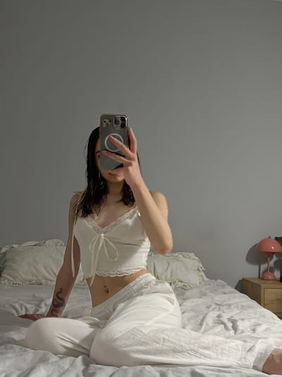 In Ternopil, Ukraine, a young woman with dark hair is taking a mirror selfie while sitting on a white, rumpled bed. She is wearing a white lace-trimmed crop top with a bow and white lounge pants. A snake tattoo is visible on her left arm. Her right hand holds a phone, obscuring her face. A pink mushroom-shaped lamp sits on a wooden nightstand to the right of the bed, against a gray wall. The room appears to be a bedroom.