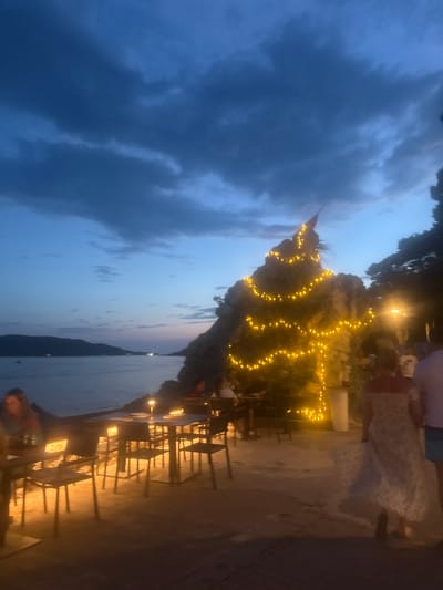 The image depicts a twilight scene at a coastal restaurant in Rafailovići, Montenegro. A rock formation, decorated with numerous yellow string lights resembling a Christmas tree, dominates the right side of the frame. The sea is visible in the background, with a silhouetted island in the distance. Several tables and chairs are set up on a paved area in front of the restaurant, some illuminated by small table lamps. People are seated at some of the tables, and others are walking along the path. The sky is dark blue with scattered clouds, indicating the transition from day to night.