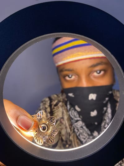 This image, taken in Ngleshi Amanfro, Ghana, depicts a close-up view of a person holding an object, framed by an illuminated black ring light. The individual's face is partially visible, with light brown eyes looking directly forward. A multi-striped knitted headband, featuring horizontal bands of pink, blue, yellow, and red, is worn on the forehead. The lower half of the face is covered by a black bandana adorned with white patterns. The person is wearing a shirt with a dark background and light brown, abstract or leaf-like patterns. A left hand, displaying short, manicured fingernails, holds a gold-colored, ornate, flower-shaped metallic object with a dark spherical center, positioning it within the ring light's glowing circumference. The background is a plain, out-of-focus bluish-gray surface.