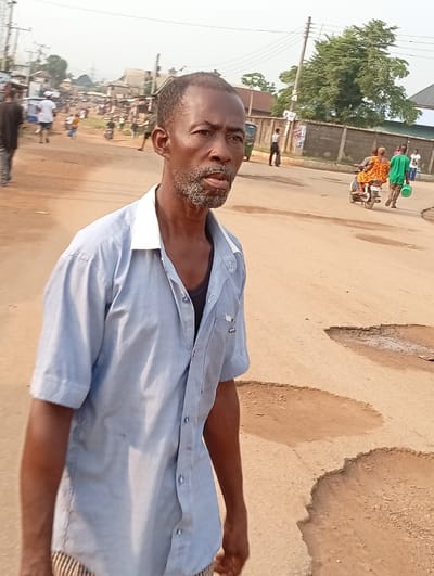 The image depicts a street scene in Umuode, Nigeria, under daylight conditions. In the foreground, a male subject, appearing middle-aged, stands slightly off-center to the left. He has short dark hair and a graying beard and mustache. He wears a light blue, short-sleeved collared shirt unbuttoned over a dark undershirt, with vertically striped lower garments visible at the bottom of the frame. His gaze is directed forward and slightly right.
The road surface is unpaved or severely degraded pavement, light brown in color, with multiple large potholes and eroded sections. In the mid-ground, several individuals are present. On the right side of the road, an individual in an orange patterned dress is seated as a passenger on a motorcycle. Nearby, another individual, wearing a green shirt, walks while carrying a green bucket. Additional pedestrians are visible further in the background. Structures lining the road include various buildings, a grey concrete wall or fence on the right, and utility poles with overhead lines. Trees are visible in the distance. The sky is bright, suggesting natural daylight.