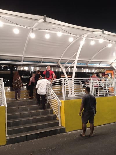 This nighttime image captures a lively outdoor or semi-outdoor dining area, likely a cafe or restaurant, in Madiun, Indonesia. The setting features a white canopy supported by white beams, adorned with string lights that illuminate the space and create a warm ambiance against the dark sky. A prominent yellow wall forms the base of the structure, leading up to a set of grey concrete stairs with white railings.

Several people are present, indicating an active scene. In the foreground, a man in a dark grey t-shirt and shorts, wearing sandals, stands with his back to the camera, holding what appears to be a cigarette. A group of individuals is seen ascending the stairs towards the main seating area. This group includes a woman in a brown patterned top and head covering, a person in a light pink top, a man in a white shirt and black peci (cap), and another man in a red t-shirt with a black backpack.

Behind the white railing, inside what looks like a seating area, at least two men are visible seated at a white table, one in a red shirt and another wearing a yellow cap. The seating area is adjacent to a dark, reflective surface resembling the side of a train car.

A significant detail is the visible text "INKA" on the side of the train-like structure. This refers to PT Industri Kereta Api, Indonesia's state-owned rolling stock manufacturer located in Madiun, strongly indicating the local context. Partially visible text to the right reads "A BUATAN INDO," likely part of "Kereta Api Buatan Indonesia" (Indonesian Made Train), further confirming the train theme. The scene suggests a casual evening gathering or dining experience at a location potentially themed around or adjacent to a train.