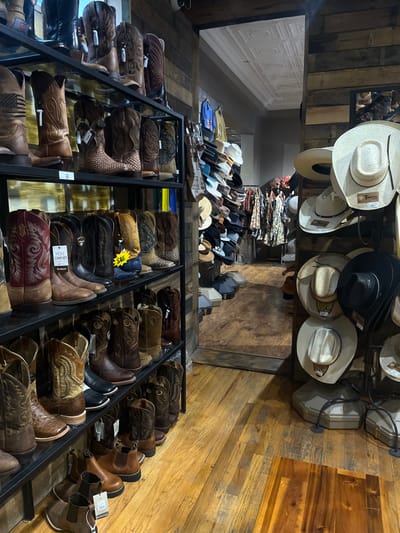 A retail space displays cowboy boots and hats. The boots are arranged on tiered metal shelves, with various styles and colors of leather. Cowboy hats are on display above, affixed to a wooden beam. A stack of fabrics rests behind the hats, near the entrance. The store has wooden floors and walls, enhancing the rustic ambiance.