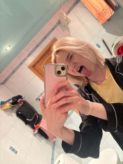 An overhead shot captures a young person in a bathroom, taking a selfie with their phone. They have short, blonde hair and are wearing a yellow t-shirt and a black robe with white trim. Their tongue is sticking out, and they are holding a pink phone with a floral pattern. The bathroom features light blue tiled walls and ceiling, a wooden door frame, and an orange towel hanging on the wall. To the left, a pile of clothes, including a black jacket and a pink item, is visible. The overall impression is one of a casual, informal moment.