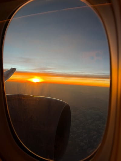 A view from an airplane window captures a dramatic sunrise. The sun is a bright orb on the horizon, casting an orange glow across the sky. Below, there's a layer of clouds. A portion of the airplane's engine and wing is visible on the right and bottom of the frame. The photo appears to be taken while flying near Taoyuan City, Taiwan, given the time of day.