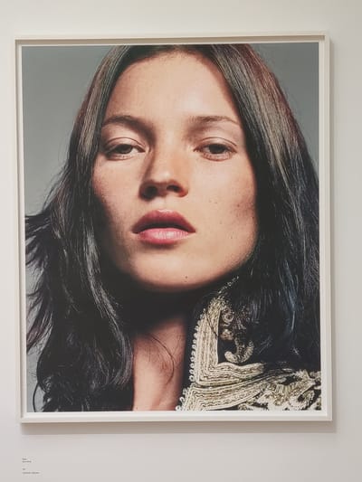 This media file is a photograph of a close-up, headshot portrait of a young woman. Her face is tilted, and her eyes are looking slightly to the side. She has dark hair that cascades around her face and shoulders. She is wearing a garment with intricate gold embroidery around the neckline. The lighting suggests it is daytime. The image is framed in white and mounted on a white wall. In the bottom right corner of the frame, there is text that reads: "Fรกtima" followed by "(Juno Mac)" and then "Fรกtima", "Juno Mac", and "Leopold van Kempen". This suggests the photograph is titled "Fรกtima" and was taken by "Juno Mac", with "Leopold van Kempen" possibly being a curator or related party. The setting appears to be an art exhibition or gallery, as indicated by the mounting and framing, and the text. The overall mood of the photograph is serene and contemplative.