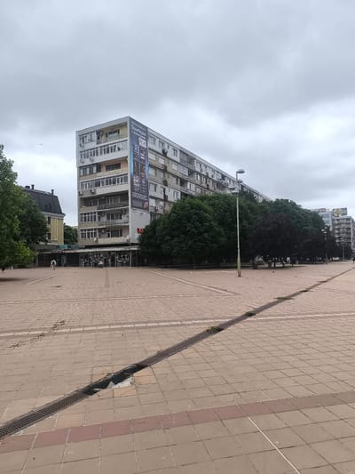 The image depicts a wide, paved public square in the city of Dobrich, Bulgaria, under an overcast sky. In the background, a tall, modern, multi-story building with numerous balconies and air conditioning units dominates the scene. A large advertisement banner is affixed to the side of this building. To the left, a smaller, older building with a yellow facade and a dark, sloped roof is visible, adjacent to trees. The square itself is paved with square tiles in shades of beige and brown, with a red band of tiles running horizontally across the foreground. A long, metal grate drainage system runs diagonally across the square, with some sections appearing damaged. Several streetlights with multiple lamps are visible, as are trees lining the perimeter of the square and between buildings. A few people are present in the square, including one person walking on the left side and a few individuals near the shops at the base of the tall building, one pushing a stroller. The overall impression is a functional urban space with a mix of modern and older architecture.