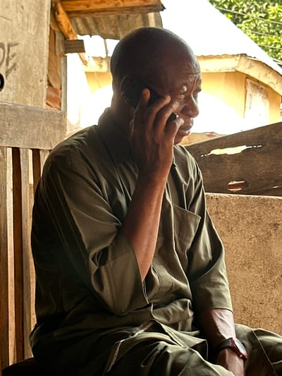An elderly Black man with dark skin and a shaved head is seated on a wooden chair, talking on a dark smartphone held to his right ear with his right hand. He is wearing a dark olive green collared shirt with the sleeves rolled up to his elbows and dark pants. A red-faced wristwatch is visible on his left wrist. The man is positioned in the foreground, with a blurred background suggesting an outdoor, possibly rural or semi-urban, setting in Mararaba, Nigeria. Behind him, vertical wooden slats form a partition. Beyond that, a weathered, rough-textured wall is visible. In the upper portion of the image, parts of a corrugated metal roof and wooden beams suggest a simple structure. The lighting is bright and casts strong shadows, indicating a sunny day. The overall atmosphere is casual and commonplace.