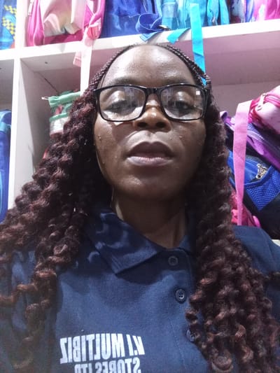 A woman with braided hair and glasses looks at the camera, her head and shoulders filling the frame. She is wearing a dark blue collared shirt with a logo that appears to read "K.I. MULTIJUM STORES LTD" in reverse. Behind her, shelves are filled with various colored backpacks. The image is a selfie taken indoors, likely in a retail setting. The lighting suggests an artificial light source.