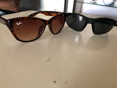 The image displays two pairs of sunglasses resting on a light-colored surface, possibly a table or countertop. The sunglasses on the left have a tortoiseshell pattern and brown gradient lenses, with the "Ray-Ban" logo clearly visible in white on the lens. The text "446 52018-137 CE" is also visible on the frame of these sunglasses. To the right, a pair of black sunglasses with dark lenses is positioned. These sunglasses have a more angular and sporty design, with textured detailing on the upper part of the frames. The background is blurred, but suggests an indoor setting with shelves and objects, possibly in a retail environment or home. The lighting suggests it is daytime, with natural light coming from an unseen source. The overall scene is static, with the sunglasses simply placed on the surface. No people are visible in the image. The provided location context, "New Stadium, Nigeria," does not have any discernible connection to the visual content of the image.
