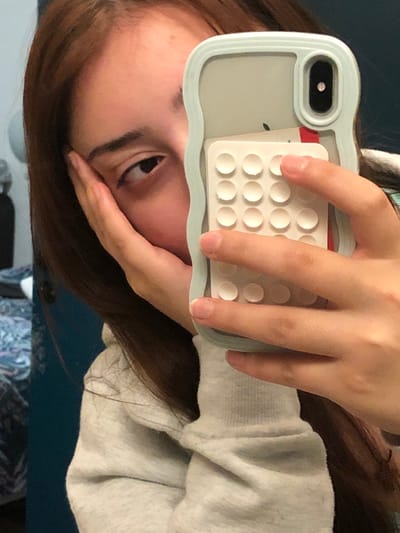A young woman with brown hair holds a light blue phone case in front of her face. The case has a white, grid-like object attached with circular bumps, resembling a pop-it toy. Her left eye is visible, looking towards the camera. Her hand partially obscures her face. She is wearing a grey sweatshirt. The background includes a blue surface and a patterned fabric. The image is rotated counterclockwise.