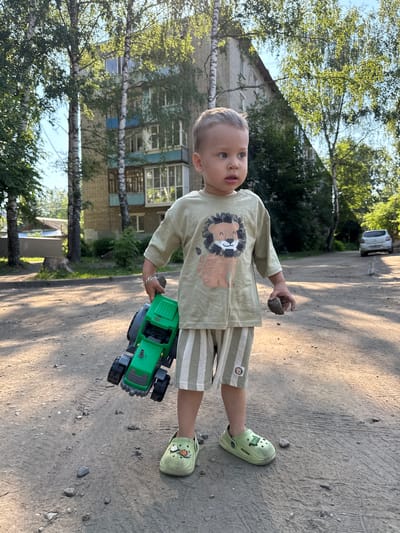 A young Caucasian boy, appearing to be around 2-3 years old, stands on a gravelly path in front of a multi-story building with a light-colored brick facade and blue trim. The boy is wearing a light green t-shirt with a cartoon lion graphic and light green and white striped shorts. He has on light green clogs with decorative charms. In his right hand, he holds a small, dark-colored rock, and in his left arm, he carries a bright green toy tractor. The setting suggests an outdoor urban environment, possibly a residential courtyard, with trees and shadows cast by the sun. A white car is visible in the background on a paved road. The boy is looking to his right, with a curious expression. The overall scene is illuminated by natural sunlight.