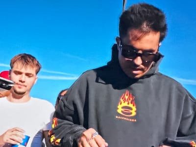The image shows two men outdoors under a clear blue sky. The man in the foreground is wearing a dark grey Balenciaga hoodie with a flame design and sunglasses. He has short dark hair and is wearing rings on his fingers. The second man is standing slightly behind and to the left, wearing a white t-shirt and holding a can of Red Bull. The location in Bragança, Portugal is unknown and not visible.