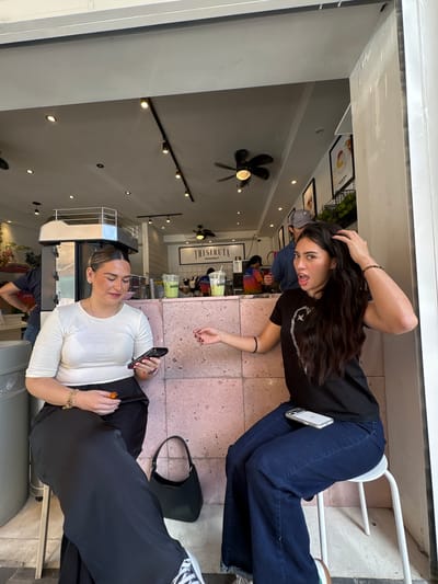 In a cafe in Puebla, Mexico, two young women are seated at a counter. The woman on the left, wearing a white top and black skirt, is looking down at her phone. She is holding a small, orange object in her right hand. A black handbag rests on the floor beside her. The woman on the right, in a black t-shirt and blue jeans, is looking forward with her mouth open, touching her hair. A smartphone is on her lap. Behind them, the cafe interior is visible, with a counter, drinks, and a menu board. The cafe appears to be modern and casual, with industrial-style lighting and a tiled wall.