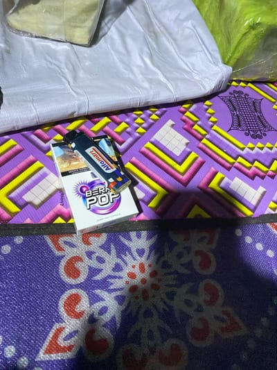 In Lamtui, Indonesia, a pack of "BERRY POP" cigarettes with a blue and white design, featuring a burst-like graphic and the text "BERRY MINT CAPSULE" is placed on a purple mat with a geometric pattern of yellow, pink, and white squares. A blue lighter with the "Alfamat" logo is resting on top of the cigarette pack. The scene is illuminated by a single light source, casting shadows. In the background, two large white and green plastic-wrapped objects are visible, one appearing to contain a light yellow substance. The mat itself has a woven texture.