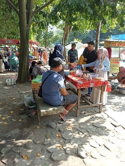 This image depicts a bustling outdoor food market or street food area in Mataram, Indonesia, characterized by several vendors and customers gathered under the shade of large trees. The ground is an uneven mix of dirt and irregular stone or concrete pavers, with scattered dry leaves.
In the foreground, a man wearing a blue short-sleeved shirt, plaid shorts, and a black baseball cap is seated on a low concrete bench, facing towards the activity at a food stall. He has a black messenger bag slung over his shoulder and is wearing flip-flops. Near his feet on the ground are a plastic crate, a clear plastic bag containing green items (possibly vegetables), a red soda can, and a straw, suggesting a casual and used environment.
The central activity revolves around a wooden table draped with a red and white patterned plastic tablecloth, serving as a food stall. A woman in a dark blue hijab stands behind the table, appearing to be a vendor as she holds a plastic bag, ready to serve. Opposite her, a customer in a light blue striped top and a light brown hijab extends her hand towards the food on the table while holding a bunch of long, thin, possibly decorative dried flowers or sticks in her other hand. A young man in a black shirt stands nearby, observing the transaction. Further in the background, another man in a striped polo shirt is also visible.
Numerous other people are present throughout the scene, some sitting at other stalls, others moving through the area, indicating a vibrant public space. Many of the women are wearing hijabs. To the right, a white food cart with red lettering clearly displays "STICK KENTANG" (Potato Sticks), pointing to the type of snacks offered. The bright sunlight filtering through the trees and creating distinct shadows suggests it is daytime, likely mid-morning or early afternoon, under clear weather conditions. The atmosphere is lively and typical of a local market.