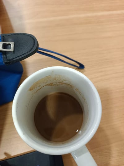 A close-up, overhead shot shows a white mug filled with a light brown liquid, likely coffee with milk. The inside rim of the mug is stained with dried residue from the drink. The mug sits on a light brown, wooden surface, suggesting a desk or table. To the left of the mug, a portion of a blue fabric pouch is visible, with a black leather flap and a silver metal clasp. A blue string or strap extends from the pouch towards the background. The scene appears to be indoors, possibly in an office or home setting, given the desk surface and the presence of a pouch. No people are visible in the frame. The lighting suggests it could be daytime, but there are no clear indicators of the time of day or weather. The overall impression is a casual, everyday moment, perhaps a break or a start to the day. No visible text is present in the image.