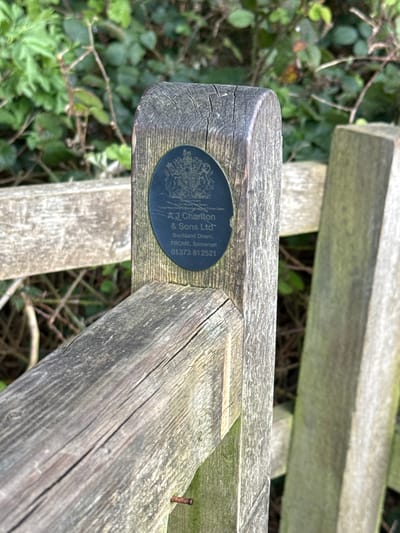 A close-up shot focuses on a weathered wooden bench, likely outdoors. The bench is constructed from thick, unpainted planks of wood, showing signs of age and weathering with cracks and a gray-brown patina. A small, circular plaque is affixed to the end of one of the planks. The plaque is dark in color with lighter text that reads "AJ Charlton & Sons Ltd, Buckland Down, FROME, Somerset, 01373 812521". The bench is situated in an outdoor environment with green foliage visible in the background, suggesting a park or garden setting. The city of Dacorum in the United Kingdom is not visible in the image.