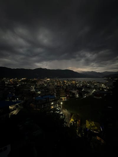 The image depicts a nighttime view of Pokhara, Nepal, likely taken from an elevated position. A mountain range divides the scene. To the left of the mountains, dark, cloudy skies dominate. To the right, the city is visible, characterized by clusters of buildings illuminated by streetlights and lights from within the structures. The darkness obscures details of the buildings. Some vegetation can be observed in the right foreground. No people are clearly visible in the image.