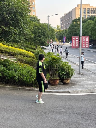 This outdoor medium shot, when correctly oriented, depicts a pathway or sidewalk alongside a road, bustling with activity, likely within a university campus or institutional area in Nan'an District, China.
In the foreground, a person stands on the sidewalk, facing away from the camera and looking towards the distant end of the path. They are wearing a black short-sleeved top with neon green accents and black shorts, along with light-colored athletic shoes. The person holds a white smartphone in their left hand.
The sidewalk is paved with grey tiles, bordered by lush green bushes and trees that create a vibrant, natural barrier between the path and the adjacent road. Several decorative potted plants are also placed along the sidewalk. Beyond these plants, the asphalt road is visible.
Further down the path, multiple individuals can be seen walking, suggesting a normal daytime flow of foot traffic. In the background, large, multi-story concrete buildings are partially visible through the foliage, typical of institutional architecture. Overhead, streetlights are present.
Two large red banners with white Chinese characters are prominently displayed on a lamppost. The text reads:
* "求真求正" (Seek truth, seek rectitude)
* "严谨严肃" (Rigorous and serious)
* "重部教风重部学风" (Emphasize departmental teaching style, emphasize departmental study style)
* "日新日进" (Daily renewal, daily progress)
* "唯实性先" (Practicality first/Prioritize practicality)
* "人才培养质量文化" (Talent cultivation quality culture)
The scene appears to be set on a clear, bright day, with soft, even lighting, possibly late morning or early afternoon. The overall impression is one of a well-maintained, active campus environment focused on education and development.