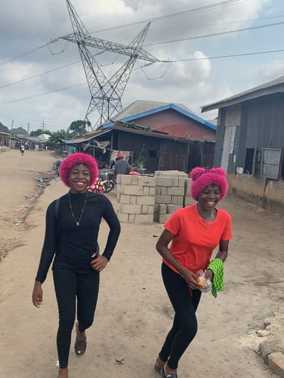 The image depicts an outdoor scene on an unpaved street in Umuode, Nigeria, under an overcast sky. In the foreground, two young females are walking towards the viewer, both smiling broadly.
The female on the left wears a black long-sleeved top, black leggings, and black slides. She has short, curly, bright pink hair (likely a wig) and a thin gold chain necklace with a small pendant. Her left hand is on her hip, and her right arm is slightly forward.
The female on the right wears an orange short-sleeved t-shirt, black leggings, and black wedge slides. She also has short, curly, bright pink hair, styled with two distinct bun-like structures on top (likely a wig). She wears a small gold chain necklace. Her hands hold a clear plastic bag containing a light-colored object, possibly food, and a folded green and white chevron-patterned cloth.
The unpaved road surface is dirt with some visible debris and loose rocks. To the right of the subjects, a stack of grey concrete blocks is present in the mid-ground. Behind the subjects, various structures line the street. These include single-story buildings with corrugated metal roofs, some with painted surfaces (e.g., a red building with a blue trim roof). A motorcycle is parked in the mid-ground near some of these structures, with a person in a red cap standing nearby. Further down the street, other individuals are visible. Dominating the upper background is a large metal electricity transmission tower/pylon with multiple power lines extending across the frame.