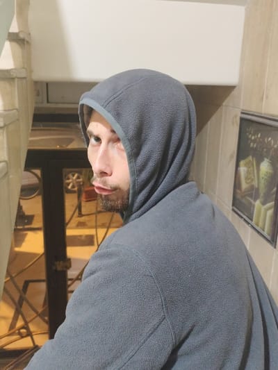 A person wearing a gray hooded fleece jacket looks over their shoulder. The person is positioned in front of a doorway leading outside, with a vehicle visible through the glass. A staircase is to the left and a framed piece of art hangs on the tiled wall to the right. The overall lighting is warm.