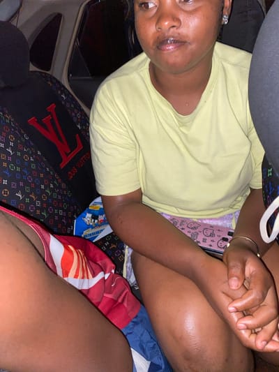 This media file shows a person, appearing to be a young Black female, sitting in a car. She is wearing a light yellow t-shirt and appears to be holding hands with someone else, though the other person is not fully visible. The background shows the interior of the car, including a black car seat with a prominent "LV" logo and a colorful pattern, possibly a seat cover or bag. There is also a pink phone case with Hello Kitty graphics visible. The person has rings on her fingers and a bracelet on her wrist. The lighting suggests it is daytime, and there are no obvious indicators of the weather. The overall mood appears relaxed and casual. The visible text includes "LOUIS VUITTON" and "Hello Kitty".