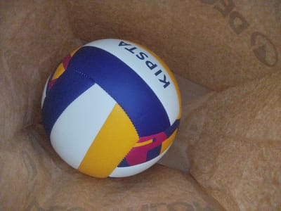 A close-up, eye-level shot captures a multi-colored volleyball positioned inside a light brown paper bag. The volleyball features panels of white, deep blue, golden yellow, and a magenta-like color with a dark outline. The brand name "KIPSTA" is visible in dark blue capital letters across one of the white panels. The paper bag's texture is rough and slightly creased, creating a soft, diffused background. Faint, dark lettering is visible on the bag's surface, suggesting branding or text from its original contents, though it is largely unreadable. The lighting appears to be soft and diffused, indicating an indoor setting or an overcast day, casting gentle shadows. No people are present in the frame, and the focus is solely on the volleyball and its immediate surroundings within the bag. The overall impression is one of simple storage or a casual arrangement of an object. The location context of Valencia, Spain, does not provide any specific visual cues within the image itself.