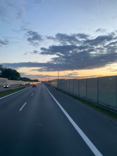 The image captures a view from a vehicle traveling on a highway near Kokotów, Poland, during twilight. The sky displays a gradient of colors, transitioning from a pale blue overhead to hues of orange and yellow near the horizon, with scattered dark clouds. The highway is paved and has clear white lane markings. Multiple vehicles are visible on the road, with their headlights on, indicating motion. On the right side of the highway, there is a tall, grey sound barrier. On the left side, there are trees and a concrete barrier. The overall scene depicts a typical highway environment at dusk.