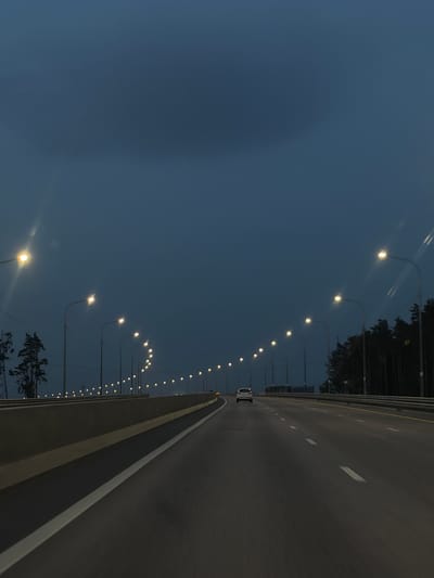 An asphalt highway with multiple lanes is depicted at dusk or night, illuminated by streetlights lining both sides of the road. The sky is a deep, dark blue, with a few lens flares from the streetlights visible in the upper left portion of the image. A single white car is traveling in the distance on the right-hand lanes. To the left of the highway, a concrete barrier separates it from what appears to be a lower level or embankment, with some trees visible on the far left. The right side of the highway is bordered by a dense line of dark trees. The image is taken from a moving vehicle, indicated by the motion blur on the road surface. The location is identified as Novomil'et, Russia, suggesting this is a road within or near that area.