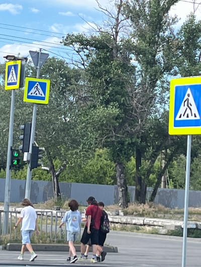 The image depicts a street scene with pedestrian crossing signs and a traffic light in Dnipro, Ukraine. Four individuals are walking across a street. The person on the far left is wearing a white t-shirt and light blue shorts. Next to them, another person is wearing a light blue t-shirt and light blue shorts. To their right, a third person is wearing a red t-shirt and black shorts, carrying a backpack. The fourth person is partially visible on the right, wearing dark clothing. The environment includes trees, a fence, and overhead power lines. The traffic light shows a green signal.