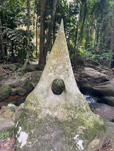 A large, weathered stone sculpture with a pointed top and a circular opening in the center stands prominently in a natural, jungle-like environment. The sculpture is covered in moss and lichen, indicating its age and exposure to the elements. It is situated beside a shallow stream or river flowing over rocks. The surrounding area is dense with tropical foliage, including trees, vines, and leafy plants. Rocks of various sizes are scattered throughout the scene, some partially submerged in the water. In the background, tall trees with hanging vines create a canopy. There is a person partially visible behind the sculpture, appearing to be interacting with the environment. The lighting suggests it is daytime, with sunlight filtering through the trees. This sculpture is located in the Samui Waterfalls area on the island of Ko Samui, Thailand.