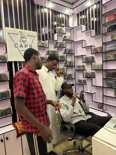 This image captures an interior scene within a cap retail establishment in Gandu Albasa, Nigeria. Three adult males are present. The shop's walls feature white, staggered display shelves showcasing numerous caps, many individually encased in clear plastic wrapping, interspersed with black and white vertical striped panels. A prominent white sign bearing the black logo "Op⁶ CAPS" and an owl illustration is affixed to the left wall. Overhead, recessed spotlights illuminate the space.
On the left, a male individual in a red and black plaid short-sleeved shirt and black track pants holds a string of light-colored prayer beads (tasbih) in his right hand. Adjacent to him, a second male, dressed in a long white traditional robe, stands looking downwards, with his right hand positioned near his chin. To the right, a third male, bearded and wearing a light grey long-sleeved shirt, black pants, and a black baseball cap, is seated in a black office chair, which is covered with clear plastic. He is actively engaged with a dark smartphone held in both hands, an earbud wire visible. A white laptop is partially visible on a counter surface to his right. A red and yellow patterned prayer mat lies partially visible on the floor in the lower left quadrant.