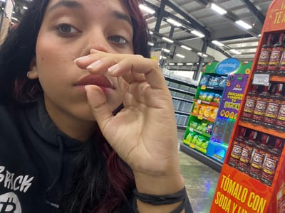 A young woman, with dark hair and a black t-shirt featuring the word "BRICK", is the primary subject of this image. She is holding her hand up to her face, partially obscuring it. The image is taken from a low angle, and the woman's body is oriented sideways. The scene appears to be a supermarket aisle, indicated by shelves filled with various products.
On the left side of the frame, there are shelves stocked with bottles of Smirnoff vodka, some of which have a red label with white text and a cartoon-like image of a person's head. Above these bottles, a large, brightly colored sign with yellow text reads "TÓMALO CON SODA Y". Further down the aisle, to the right of the vodka, are shelves filled with snack bags, displaying colorful graphics and text in Spanish. One sign reads "Snacks" in large, colorful letters. Another sign, a blue oval with white text, says "Crustissimo".
The background reveals a well-lit retail environment with rows of fluorescent lights on the ceiling. The overall lighting suggests an indoor setting, likely during the daytime, given the brightness. There are no overt indications of the weather or time of day beyond the artificial lighting. The woman's expression is not fully visible due to her hand, but the visible parts of her face do not convey strong emotions. The presence of the retail environment and the product displays suggests a shopping or casual visit to a store. The text "Pampatar, Venezuela" provides the geographical context for the scene.