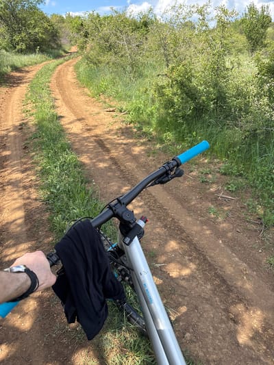 The image presents a first-person perspective of a person holding the handlebars of a mountain bike. The bike is on a dirt trail, suggesting a rural or outdoor setting. Vegetation, including trees and grass, lines the trail on both sides. The bike frame is silver, and the handlebar grips are blue. A black garment is draped over the handlebars. The person's arm and a watch are visible in the foreground. The location is not definitively identified as Katina, Bulgaria; that information is not discernible from the visual content.