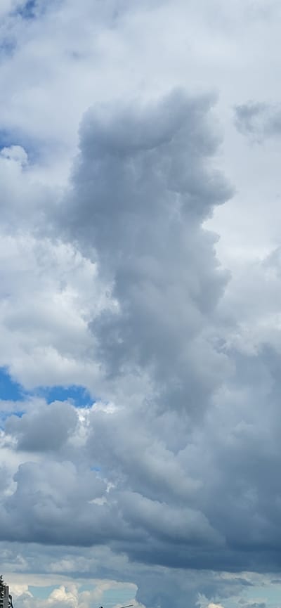 The image displays a cloudscape with a prominent, large, grey cumulonimbus cloud dominating the center. The cloud has a textured appearance with darker areas indicating depth and potential for rain. Flashes of blue sky are visible through gaps in the clouds, particularly in the upper left and lower middle sections. Lighter, fluffier clouds are scattered around the main formation. The bottom of the image shows the tops of some buildings and structures, suggesting an urban or semi-urban environment. Based on the provided context, this scene is located in Bulgakovo, Russia, and depicts a cloudy sky above the city.