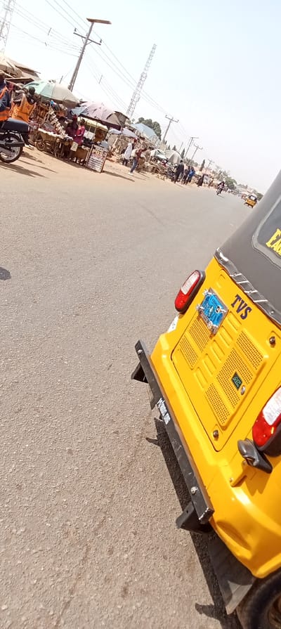 The image captures a street scene in Rido, Kaduna State, Nigeria, viewed from the rear-right perspective of a yellow TVS-branded auto-rickshaw. The auto-rickshaw features a black roof, red taillights, a black rear bumper, and a blue license plate with "KADUNA" visible. The vehicle is on an asphalt road surface.
To the left, a lively market lines the roadside, comprising multiple vendor stalls shaded by various fabric and plastic awnings, including light green, dark grey, and blue. Goods, consistent with general market merchandise and electronics, are displayed on tables and carts. A prominent "POS" (Point of Sale) service sign is visible on a standing placard. Multiple individuals are present within the market area, dressed in varied casual clothing, engaged in buying, selling, or walking. A black motorcycle is partially visible parked near the market stalls on the far left.
In the background, utility poles with power lines and a tall lattice telecommunications tower are visible against a clear, bright sky. Another yellow auto-rickshaw can be seen further down the road, receding into the distance.
