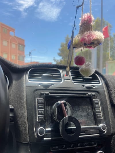 Here is a description of the image:

The image shows the interior dashboard of a car, focusing on the center console. An aftermarket car phone holder is affixed, and a car key is attached to it. Hanging from the rearview mirror is a decoration consisting of yarn and feathers. Outside of the vehicle, the sky is clear and buildings are visible, suggesting it is daytime. A partial view of an orange-colored building is visible through the windshield. Based on the building style, and the items present in the car, it is highly probable the location of this image is Ouaouizert, Morocco, since those kinds of decorations are common there.