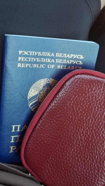 A close-up shot shows a portion of a blue Belarusian passport and a maroon leather coin purse or small wallet. The passport has the text "РЭСПУБЛІКА БЕЛАРУСЬ", "РЭСПУБЛІКА БЕЛАРУСЬ", and "REPUBLIC OF BELARUS" printed in gold lettering. A golden emblem, likely the national emblem of Belarus, is visible on the passport. The coin purse is positioned in the foreground, obscuring part of the passport, and its textured leather surface is clearly visible. The background consists of a dark, textured fabric. The scene appears to be laid out on a flat surface.