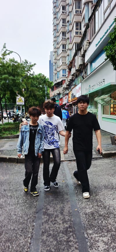 Three individuals are positioned on a sidewalk in Jiulongpo District, China, with buildings and greenery visible in the background. The individual at the top wears a black shirt and pants, sunglasses, and white shoes, and is holding a cell phone in their left hand and the arm of the individual in the middle. The middle person has on a white t-shirt, blue and white print, black pants, and black and white shoes. The bottom individual sports a denim jacket, black t-shirt, black pants, and yellow sneakers.
