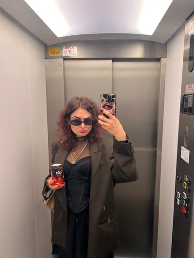 A female individual is depicted in an elevator in the city of Picassent, Spain, taking a selfie using a smartphone. She has dark, wavy, shoulder-length hair and wears black oval sunglasses, red lipstick, and red nail polish on her left hand. Her attire includes a black sheer high-neck top, a black corset-style top, an oversized dark olive green blazer, and black drawstring pants. She accessorizes with multiple gold-toned layered necklaces and a gold-toned ring on her left ring finger. In her left hand, she holds a black and red "Coca-Cola Zero" can. Her right hand holds a smartphone, equipped with a case featuring a stylized eye pattern, facing the reflective surface to capture the image. The elevator interior consists of silver-colored metal doors and light gray side walls. A white illuminated strip light is visible on the ceiling. Reflections on the elevator surfaces display reversed text, including "2GTA" and "ATES," alongside other unidentifiable characters, a yellow rectangular information sign, and a control panel with illuminated buttons.