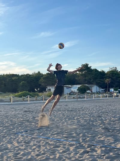 A young male volleyball player is captured mid-air during a jump on a sandy beach, serving or spiking a ball. The player is wearing a dark t-shirt with a green accent and black shorts. His body is arched backward, with one arm extended upwards towards the ball, which is visible in the upper center of the frame. His legs are bent, and his feet are off the ground, with sand being kicked up from his jump. The beach appears to be a volleyball court, indicated by a blue line visible in the sand. In the background, there is a line of trees, a small building with "TOILET" signage, and a clear blue sky with sparse white clouds. The setting suggests an outdoor recreational area, possibly a beach resort, which aligns with the known locations in Pistula, Montenegro, a region known for its coastal attractions. The lighting indicates it is likely late afternoon or early evening, with the sun casting soft light.