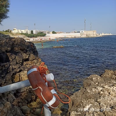 The image depicts a scenic coastal view with a clear blue sky and calm sea. The foreground is dominated by rocky terrain and a life preserver with orange rope, attached to a white pole. In the background, a beach lined with buildings and trees stretches along the coastline, with a few people visible on the beach and in the water, suggesting a leisurely day. The presence of construction cranes in the distance indicates ongoing development in the area. The lighting suggests it is daytime, possibly late afternoon given the warm tones on the rocks. Visible text on the image reads "23.06.2026 09:01". The overall atmosphere is tranquil and typical of a summer day by the sea in a populated coastal town.